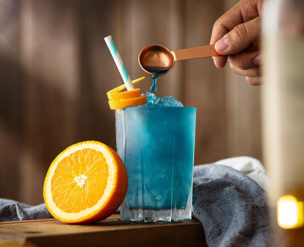 A person pours a vibrant blue liquid into a glass, garnished with a fresh orange slice on the rim.