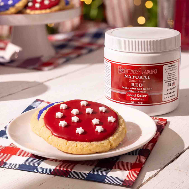 White tub labeled, "Natural Ultra Pure Red Made with Red Radish & Red Tomato Food Color Powder" next to a plate with a red colored cookie on it. 