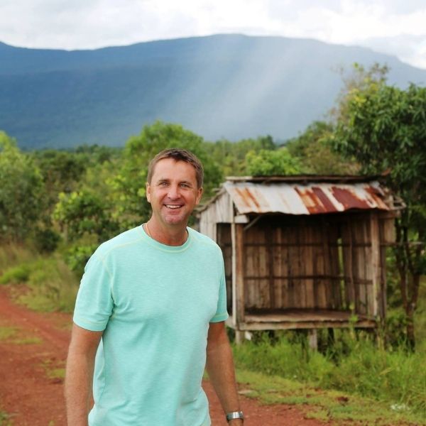 Man in a light blue shirt standing in a rural setting with mountains in the background