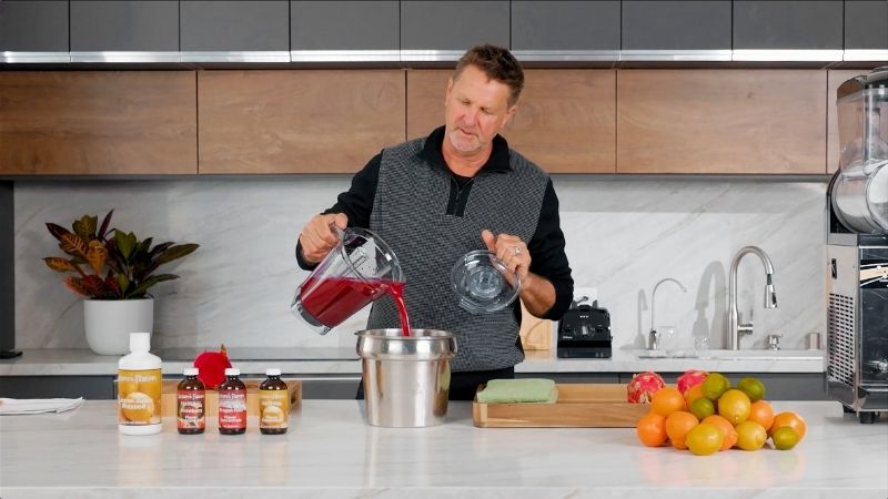 Man in a kitchen pouring red liquid into a silver container with various items on the counter.