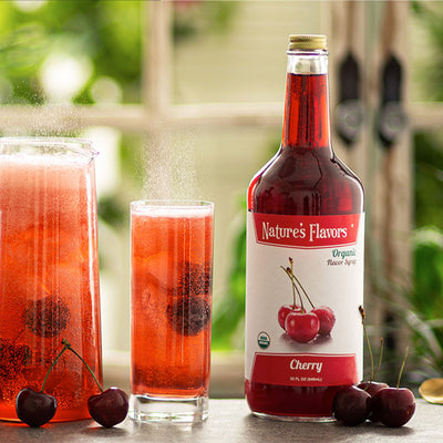 A red bottle labeled, "Organic Flavor Syrup Cherry" next to cherries and glasses of red soda with cherries in them. 