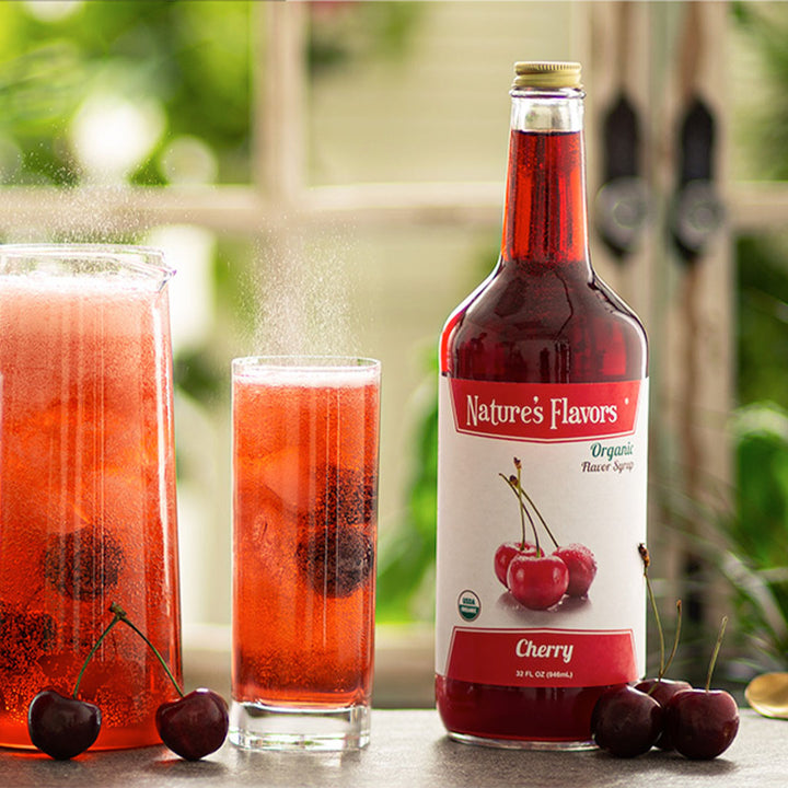 A red bottle labeled, "Organic Flavor Syrup Cherry" next to cherries and glasses of red soda with cherries in them. 