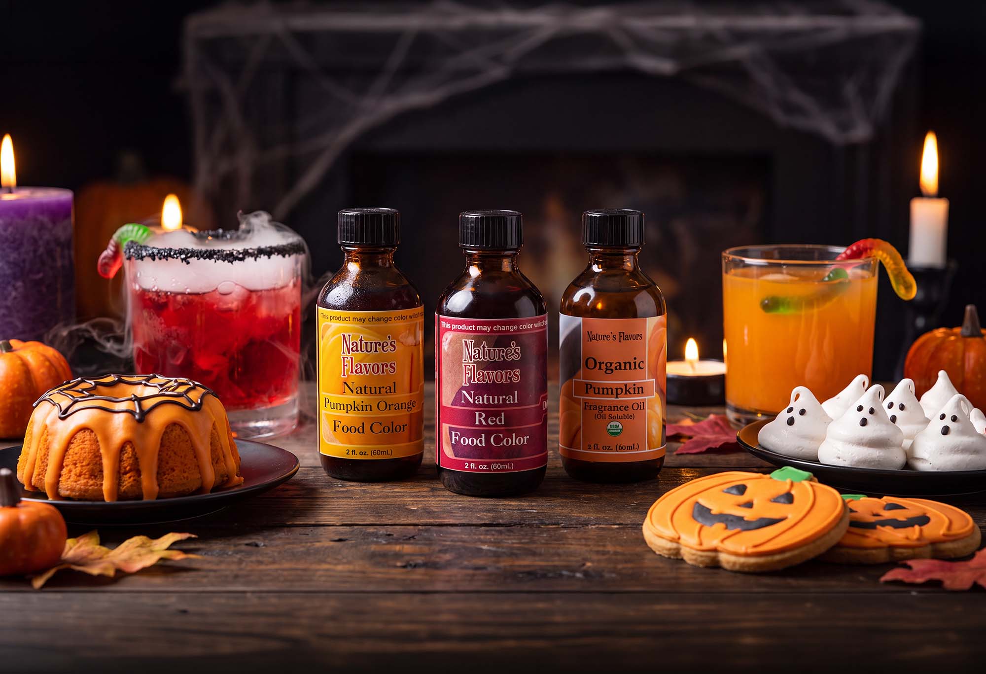 Four bottles of Nature's Flavors Natural Food Color and flavors arranged on a rustic wooden table with Halloween decorations, including candles, a pumpkin, ghost cookies, and a decorated cake.