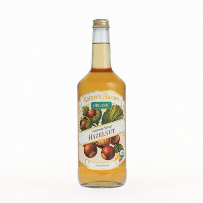 A bottle of Nature's Flavors Organic Gourmet Syrup Hazelnut, filled with golden syrup and featuring a white label with illustrations of hazelnuts and green leaves, set against a plain white background.