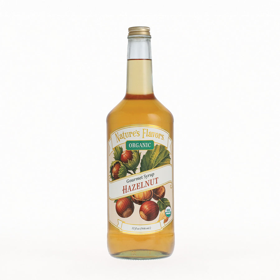 A bottle of Nature's Flavors Organic Gourmet Syrup Hazelnut, filled with golden syrup and featuring a white label with illustrations of hazelnuts and green leaves, set against a plain white background.