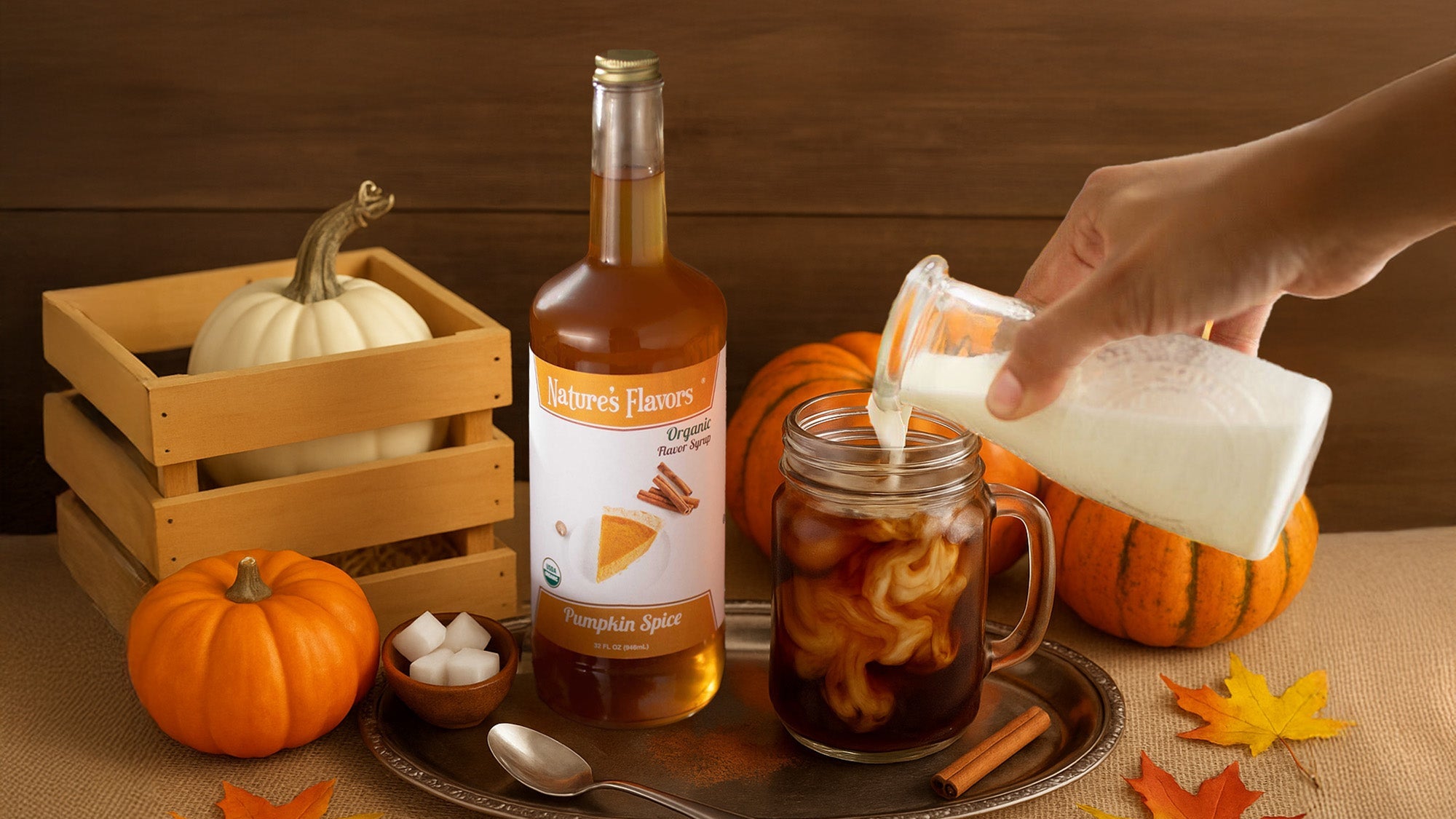 Person pouring milk into a glass of pumpkin spice latte with pumpkins and autumn leaves on a wooden surface.