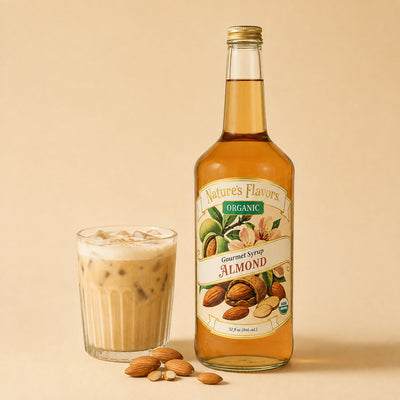 A bottle of Nature's Flavors Organic Gourmet Syrup Almond stands next to a glass of iced coffee with foam, with a few almonds scattered on a warm beige background.