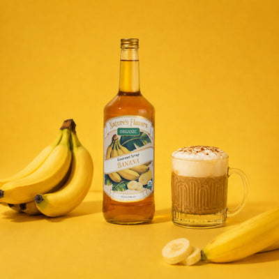 A bottle of Nature's Flavors Organic Banana Syrup stands on a yellow background next to a glass mug filled with a banana-flavored drink topped with banana cold foam, with bananas placed around the scene.