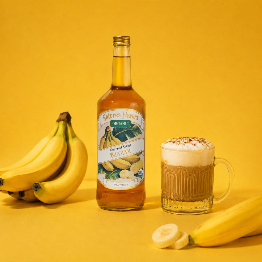 A bottle of Nature's Flavors Organic Banana Syrup stands on a yellow background next to a glass mug filled with a banana-flavored drink topped with banana cold foam, with bananas placed around the scene.