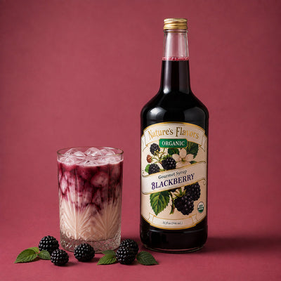 A bottle of Nature's Flavors Organic Gourmet Syrup Blackberry stands next to a glass of milk tea with berry syrup layered on top, with fresh blackberries and mint leaves on a dusty rose background.