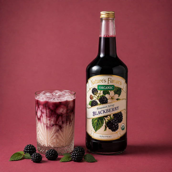 A bottle of Nature's Flavors Organic Gourmet Syrup Blackberry stands next to a glass of milk tea with berry syrup layered on top, with fresh blackberries and mint leaves on a dusty rose background.
