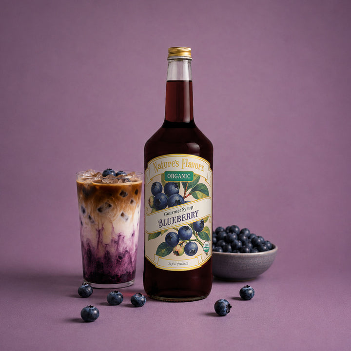 A bottle of Nature's Flavors Organic Blueberry Syrup stands next to a glass of iced coffee with blueberry syrup, with a small bowl of fresh blueberries on a purple background.