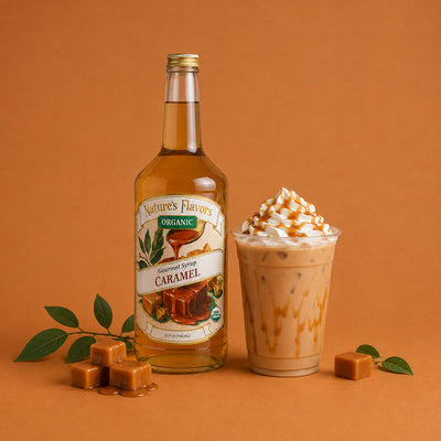 A bottle of Nature's Flavors Organic Caramel Syrup stands next to a tall plastic cup filled with an iced caramel-latte topped with whipped cream and caramel drizzle, set on a warm orange-brown background with caramel cubes and green leaves.