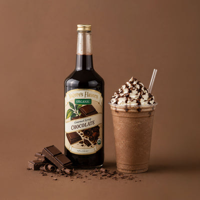 A bottle of Nature's Flavors Organic Chocolate Syrup stands next to a chocolate milkshake topped with whipped cream and drizzles, with chocolate pieces scattered on a brown studio background.
