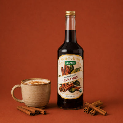 A bottle of Nature's Flavors Organic Cinnamon Gourmet Syrup stands on a warm rust-brown background, styled with a ceramic mug of cinnamon hot cocoa and scattered cinnamon sticks.