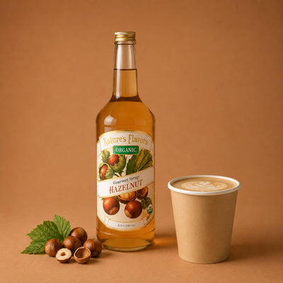 A bottle of Nature's Flavors Organic Hazelnut Gourmet Syrup stands on a warm brown background next to a paper coffee cup and a few hazelnuts with a green leaf.