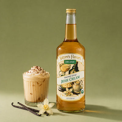 A bottle of Nature's Flavors Organic Irish Cream Gourmet Syrup stands next to a prepared iced coffee drink topped with whipped cream and cocoa powder, with vanilla beans and a flower on a muted green background.