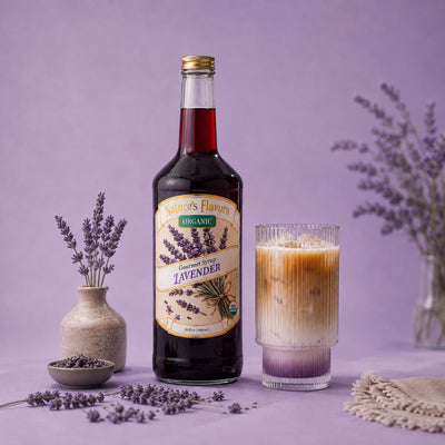 A bottle of Nature's Flavors Organic Gourmet Syrup Lavender stands next to a glass of iced coffee with lavender syrup, styled with lavender sprigs and a small vase on a purple background.