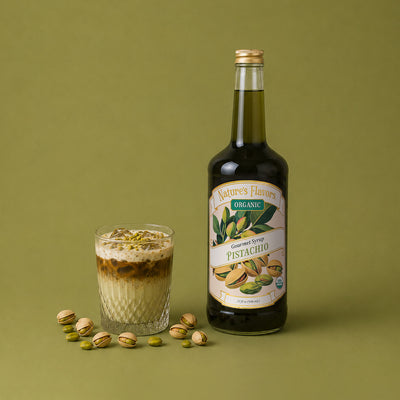 A bottle of Nature's Flavors Organic Pistachio Syrup stands next to a glass of iced pistachio latte topped with crushed pistachios, with scattered whole pistachios on a solid olive green background.
