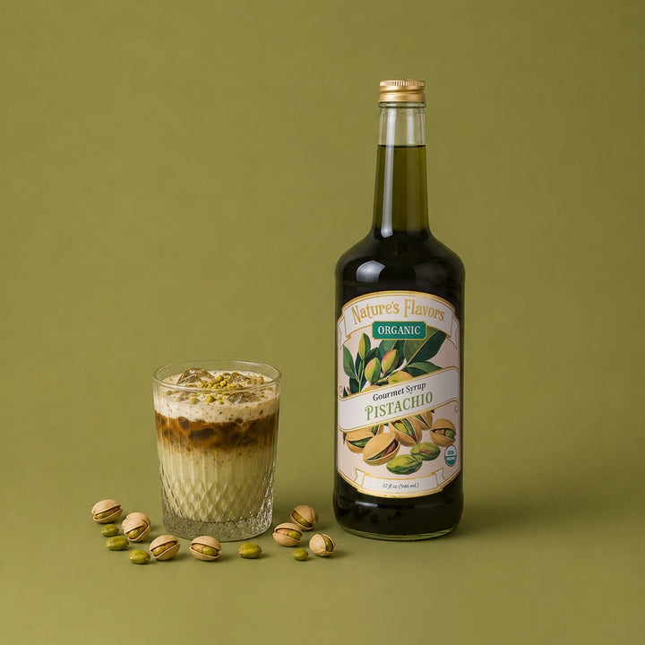 A bottle of Nature's Flavors Organic Pistachio Syrup stands next to a glass of iced pistachio latte topped with crushed pistachios, with scattered whole pistachios on a solid olive green background.