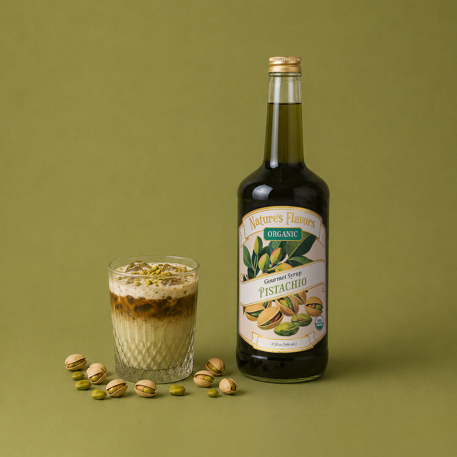 A bottle of Nature's Flavors Organic Pistachio Syrup stands next to a glass of iced pistachio latte topped with crushed pistachios, with scattered whole pistachios on a solid olive green background.