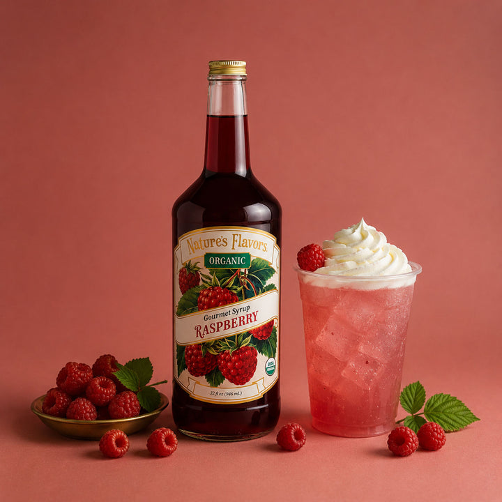 A bottle of Nature's Flavors Organic Gourmet Syrup Raspberry stands on a pink studio background, styled with a pink iced drink topped with whipped cream and raspberries, plus loose raspberries and leaves.