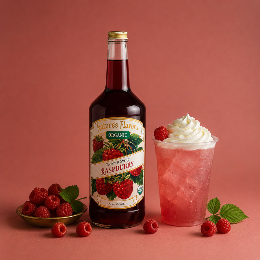A bottle of Nature's Flavors Organic Gourmet Syrup Raspberry stands on a pink studio background, styled with a pink iced drink topped with whipped cream and raspberries, plus loose raspberries and leaves.