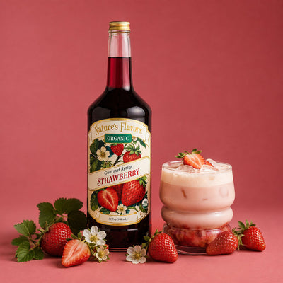A bottle of Nature's Flavors Organic Strawberry Gourmet Syrup stands next to a pink strawberry drink in a glass, surrounded by fresh strawberries, strawberry leaves, and flowers on a solid pink background.