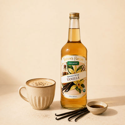 A bottle of Nature's Flavors Organic Vanilla Gourmet Syrup stands on a beige surface next to a ceramic mug filled with a frothy coffee drink, vanilla beans, and a small dish of vanilla extract.