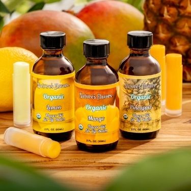 Three bottles of Nature's Flavors Organic Mango lip balm on a wooden surface with fruits in the background.