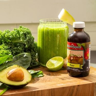 Green smoothie in a glass jar with ingredients including an avocado, lime, and a bottle of juice concentrate on a wooden surface.