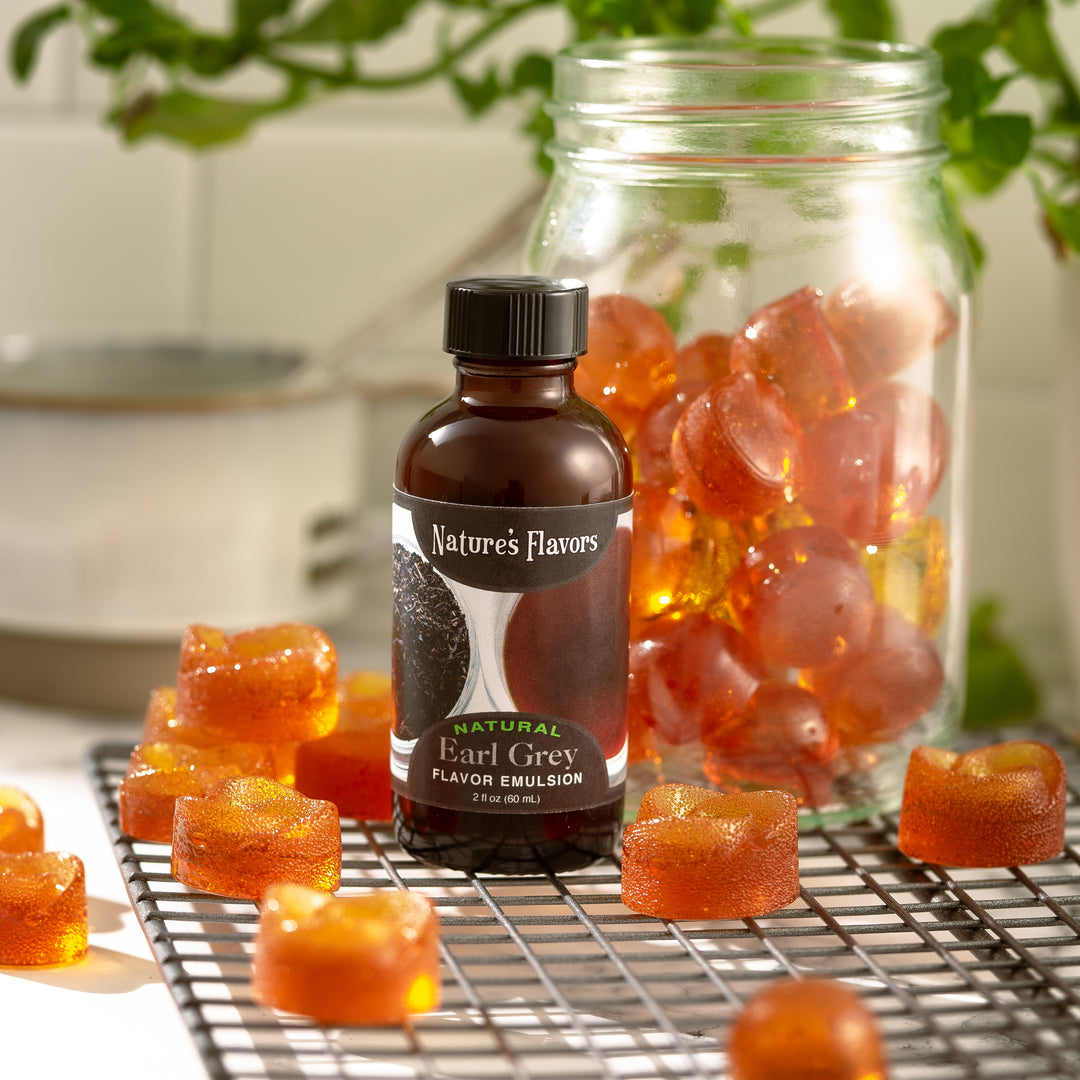 Bright orange hard candies are in a jar next to a glass bottle of Nature's Flavors Earl Grey Flavor Emulsion, creating a colorful and inviting candy scene.