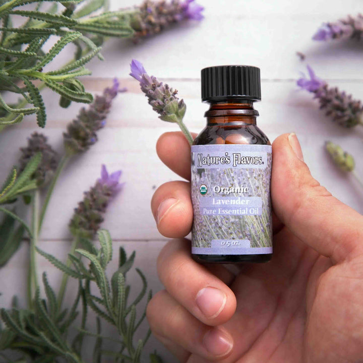 Hand holding a bottle of Nature's Flavors Organic Lavender Pure Essential Oil with lavender flowers in the background.