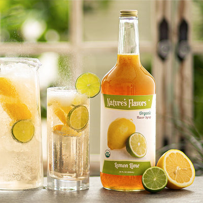 bottle labeled, "Organic Flavor Syrup Lemon Lime" next to a sliced lemon and lime and two glasses of bubbling clear soda. 