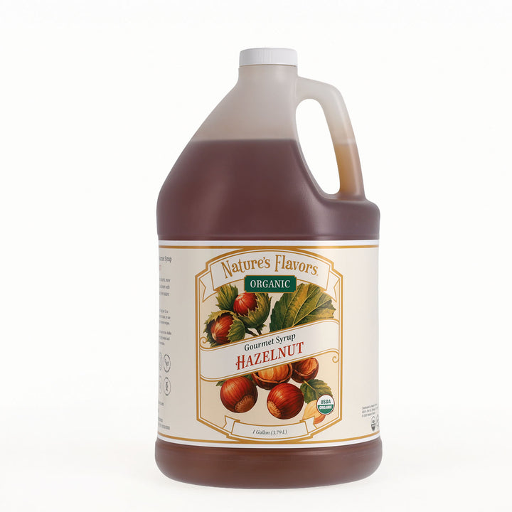 A one-gallon plastic jug of Nature's Flavors Organic Hazelnut Gourmet Syrup, filled with dark brown syrup and featuring a cream-colored label with illustrations of hazelnuts and green leaves, set against a plain white background.