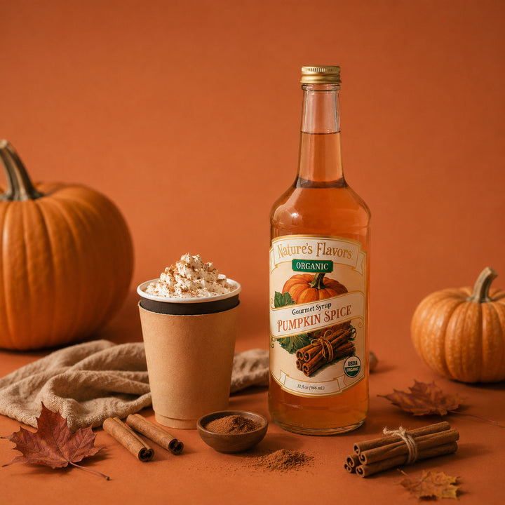 A bottle of Nature's Flavors Organic Pumpkin Spice syrup stands on a warm orange surface beside a pumpkin spice latte topped with whipped cream and cocoa, with pumpkins, cinnamon sticks, a small bowl of ground spice, and autumn leaves around it.