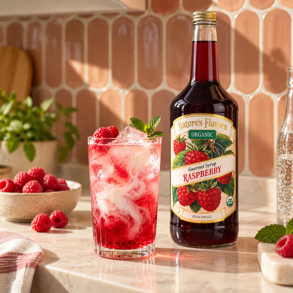 A bottle of Nature's Flavors Organic Raspberry Syrup stands on a kitchen counter beside a tall glass filled with iced raspberry drink and fresh raspberries. The background shows a peach tile wall, a bowl of raspberries, and a potted plant.