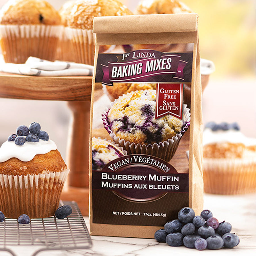 Package labeled, "Gluten Free vegan Blueberry muffin" standing next to a batch of blueberries and a blueberry muffin. 