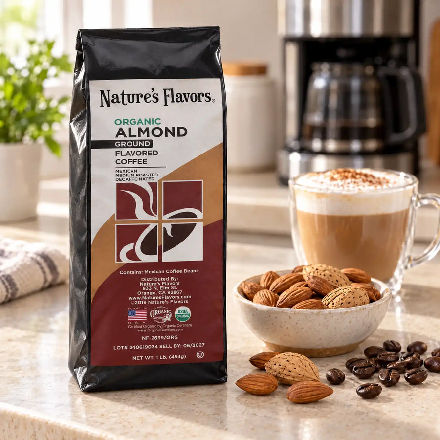 Nature’s Flavors Organic Decaf Almond Flavored Coffee Beans, 1 lb. Ground, in a black labeled bag beside almonds, whole roasted coffee beans and a filled coffee drink in a bright coffee scene.