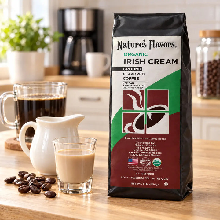 Nature’s Flavors Organic Decaf Irish Cream Flavored Coffee Beans, 1 lb. Ground, in a black labeled bag beside Irish cream and a cream pitcher, whole roasted coffee beans and a filled coffee drink in a bright coffee scene.