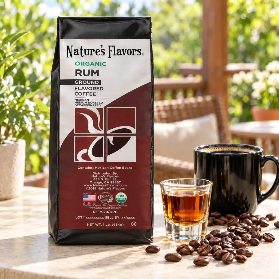 Nature’s Flavors Organic Decaf Rum Flavored Coffee Beans, 1 lb. Ground, in a black labeled bag beside a small glass of rum, whole roasted coffee beans and a filled coffee drink in a bright coffee scene.