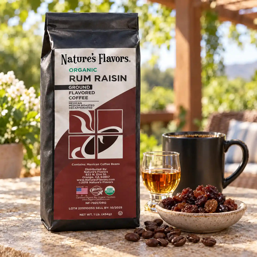 Nature’s Flavors Organic Decaf Rum Raisin Flavored Coffee Beans, 1 lb. Ground, in a black labeled bag beside raisins and a small glass of rum, whole roasted coffee beans and a filled coffee drink in a bright coffee scene.