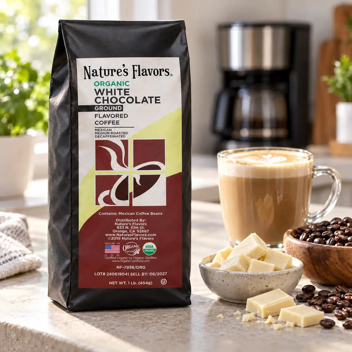 Nature’s Flavors Organic Decaf White Chocolate Flavored Coffee Beans, 1 lb. Ground, in a black labeled bag beside white chocolate, whole roasted coffee beans and a filled coffee drink in a bright coffee scene.