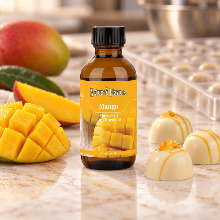 Nature's Flavors Mango Flavor Oil for Chocolate bottle beside cut mango and white chocolate pralines on a marble workbench.