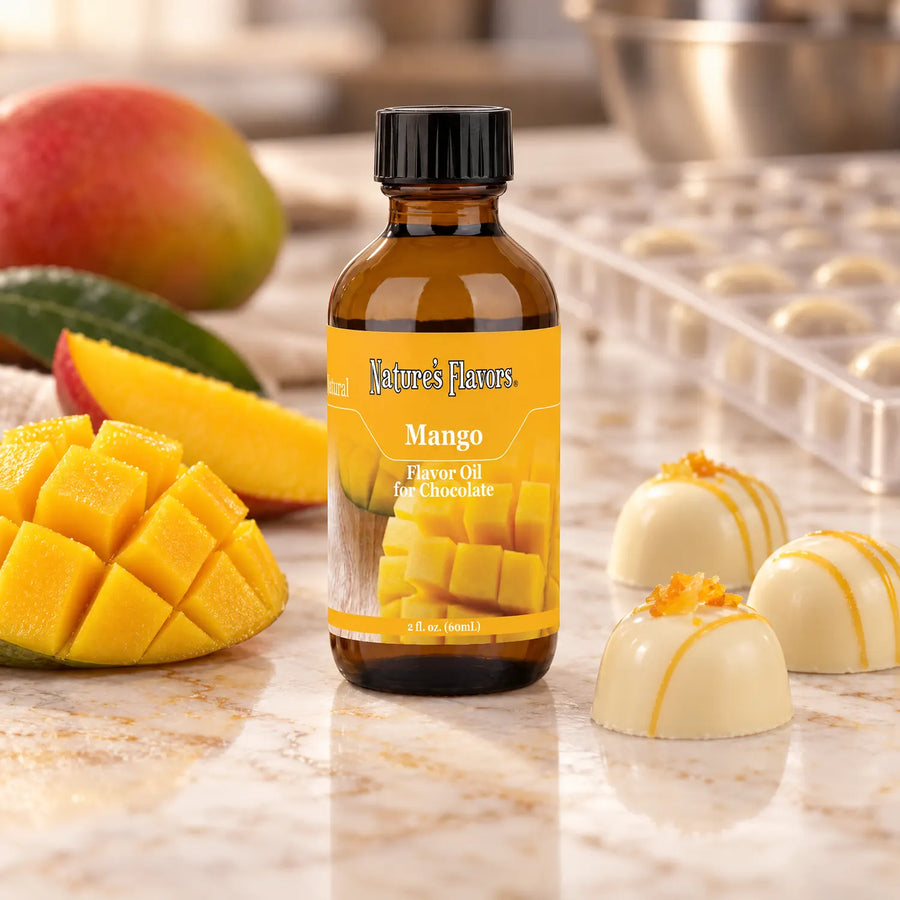 Nature's Flavors Mango Flavor Oil for Chocolate bottle beside cut mango and white chocolate pralines on a marble workbench.