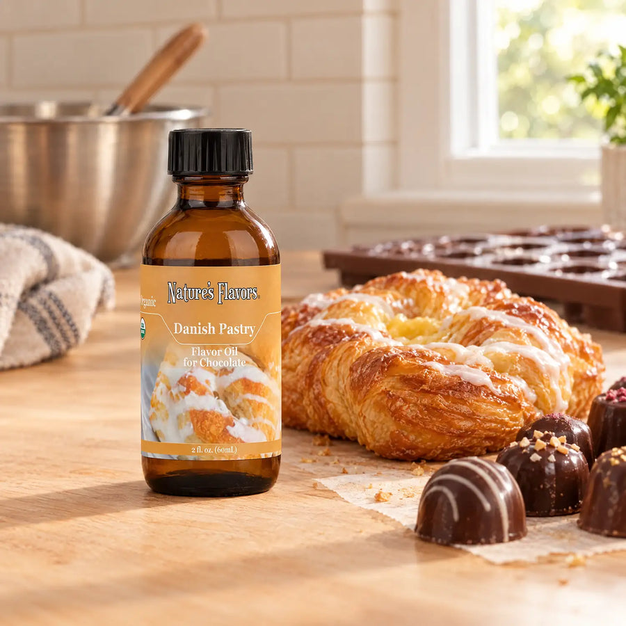 Nature's Flavors Organic Danish Pastry Flavor Oil For Chocolate, 2-ounce bottle, with a Danish pastry and chocolate bonbons in a working kitchen.