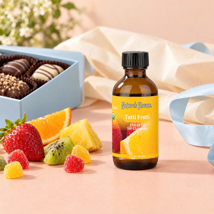 Nature's Flavors Organic Tutti Frutti Flavor Oil For Chocolate, 2-ounce bottle, with mixed fruit and boxed chocolates on a gift table.