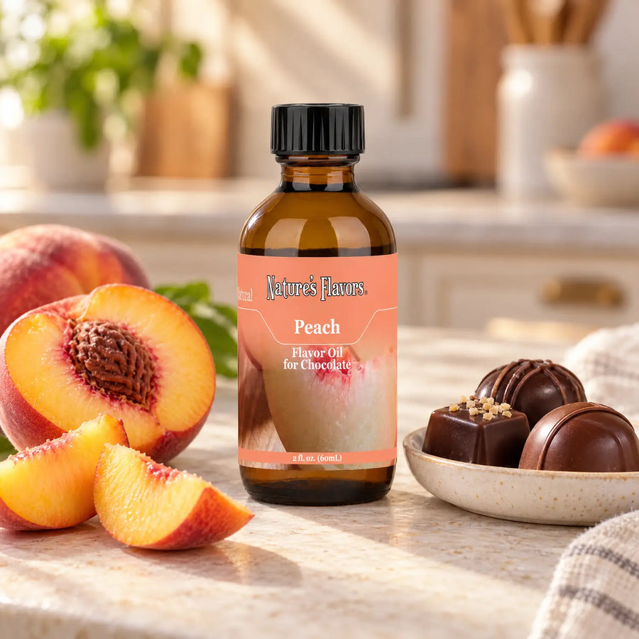 Nature's Flavors Peach Flavor Oil for Chocolate bottle beside peach halves and slices and chocolate bonbons on a sunlit kitchen counter.