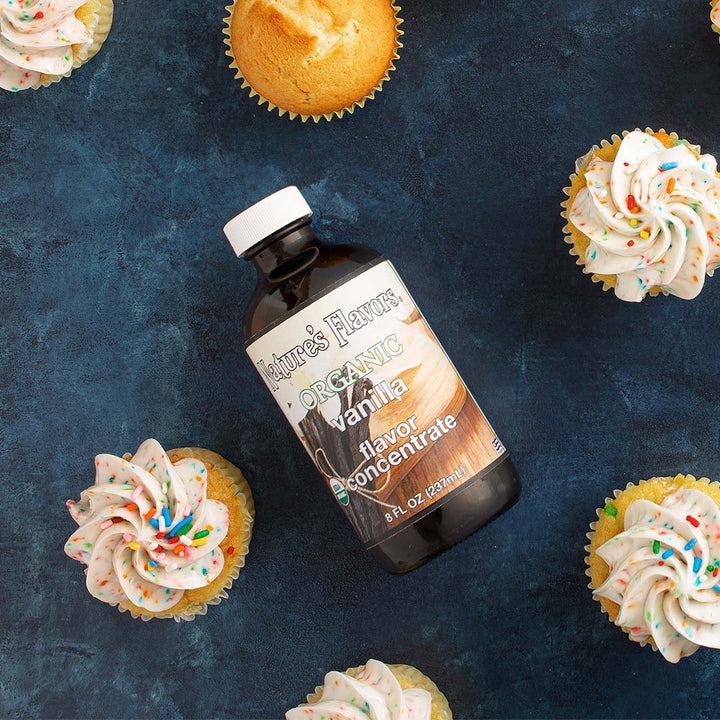 Bottle labeled, "Organic Vanilla Flavor Concentrate" lying down next to a group of cupcakes. 