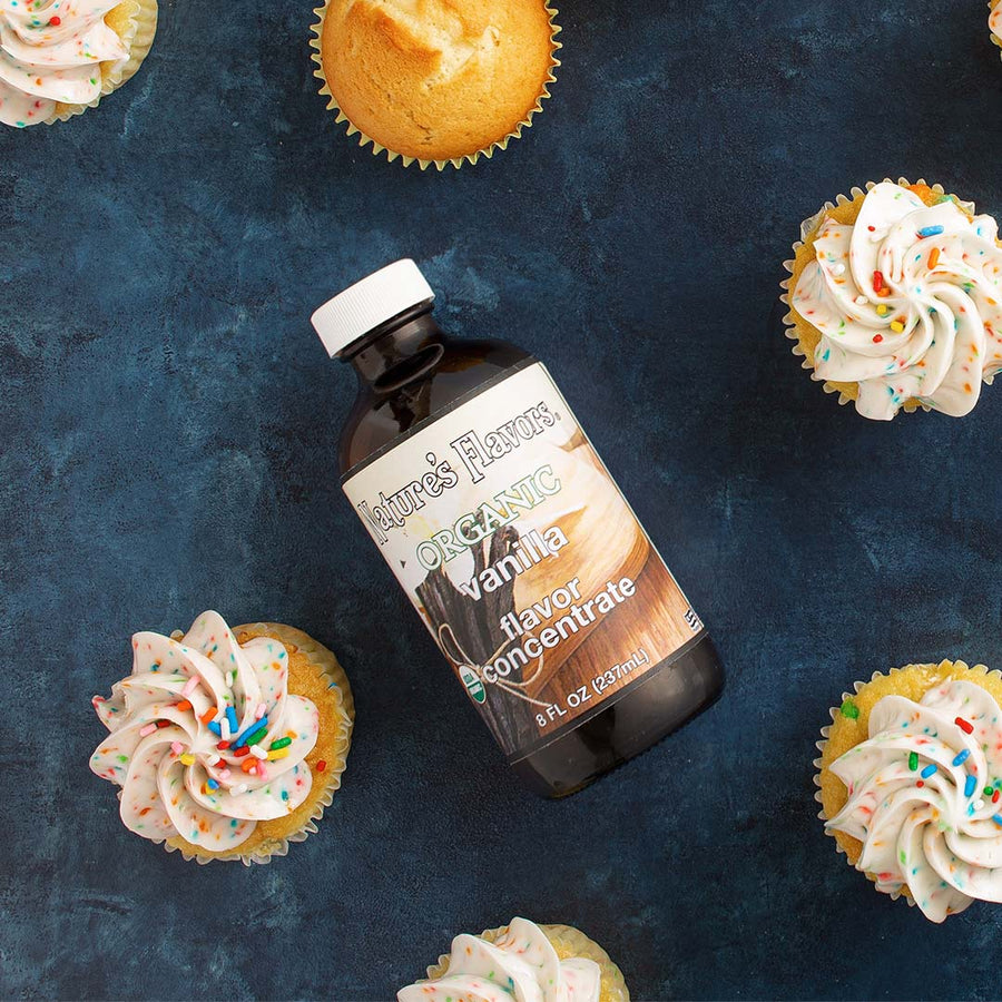 Bottle labeled, "Organic Vanilla Flavor Concentrate" lying down next to a group of cupcakes. 
